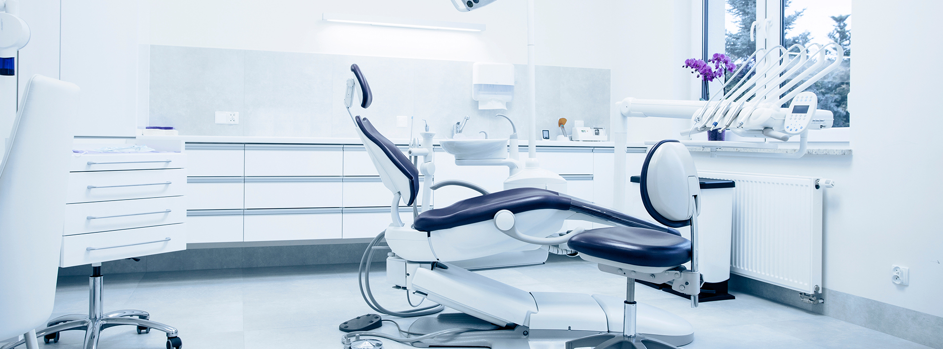 The image shows a modern dental office interior with various pieces of equipment and furniture, including a dental chair, workstations, and cabinets.