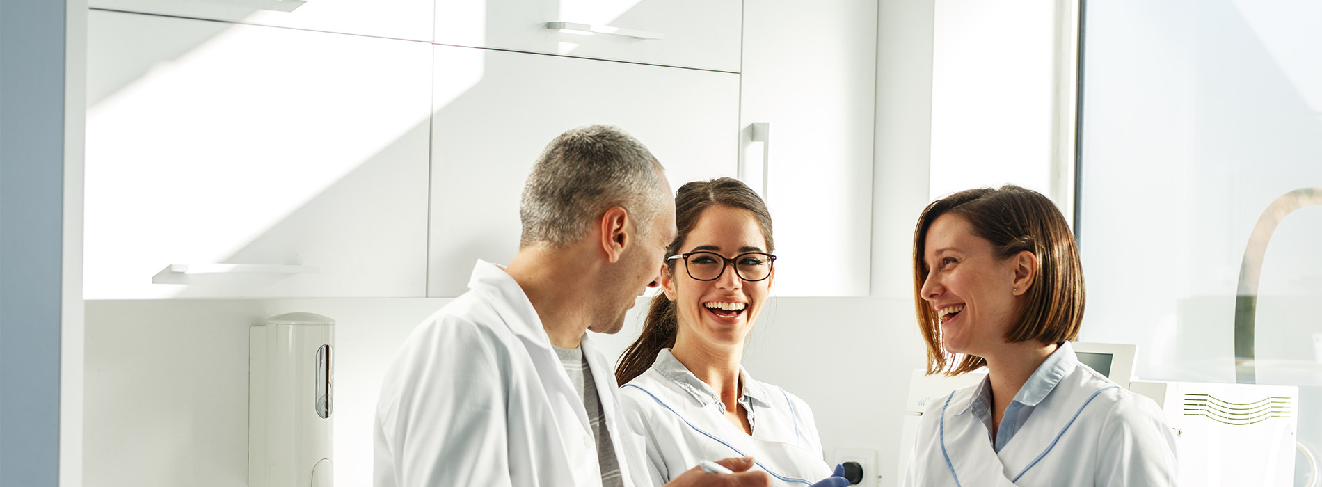 The image shows a group of three individuals, two seated and one standing, in a professional setting with medical attire, smiling at the camera.