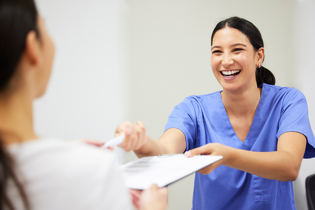 The image shows a female nurse in a white uniform, smiling at the camera, handing a document to a patient who is seated and appears to be receiving the paperwork.