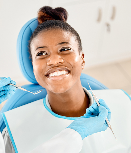 The image shows a smiling woman in a dental chair, receiving dental care with a dental hygienist holding instruments and wearing protective gloves.