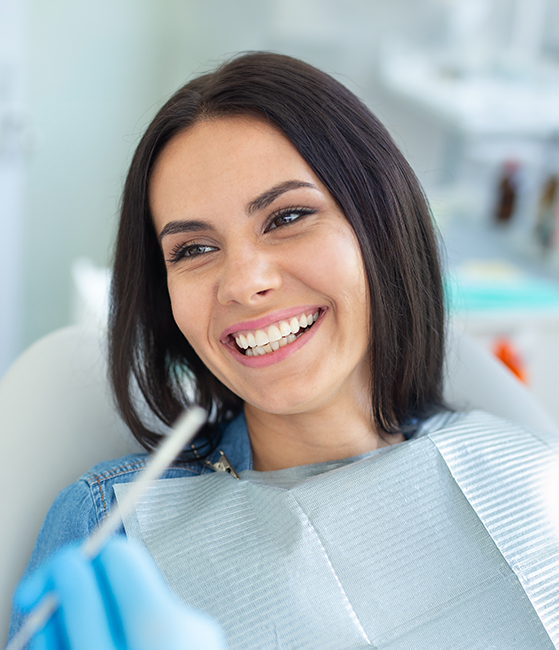 A woman is smiling while sitting in a dental chair, with her teeth being cleaned by a hygienist who is holding a dental tool.