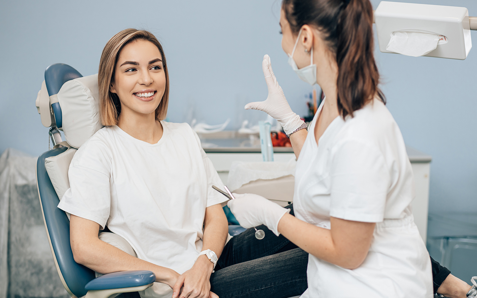 The image shows a dental office setting with a woman sitting in a dentist s chair, receiving treatment, while another woman, presumably the dentist, is standing behind her, wearing protective gear and holding a dental instrument.