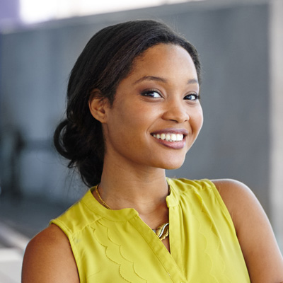 A young woman with a radiant smile, wearing a yellow top and standing in front of a building.