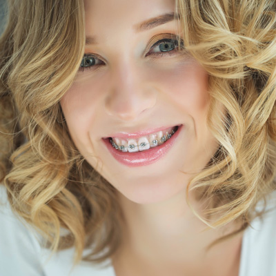 A woman with a radiant smile and straight, white teeth is the focal point of this image. Her hair is styled in loose curls that fall gently around her face. She has a fair complexion and her makeup includes subtle eyeliner and mascara, enhancing her natural beauty. The background is minimalistic, with soft lighting that highlights her features without casting harsh shadows.