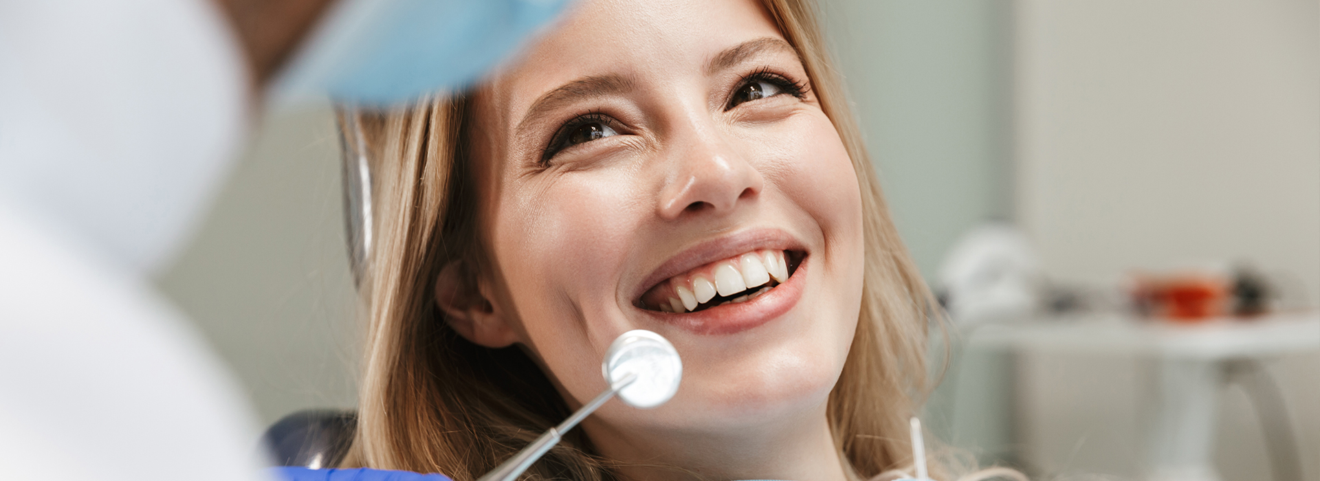 A smiling woman with blonde hair, wearing a white lab coat, is seated in front of a medical professional who appears to be examining her teeth.