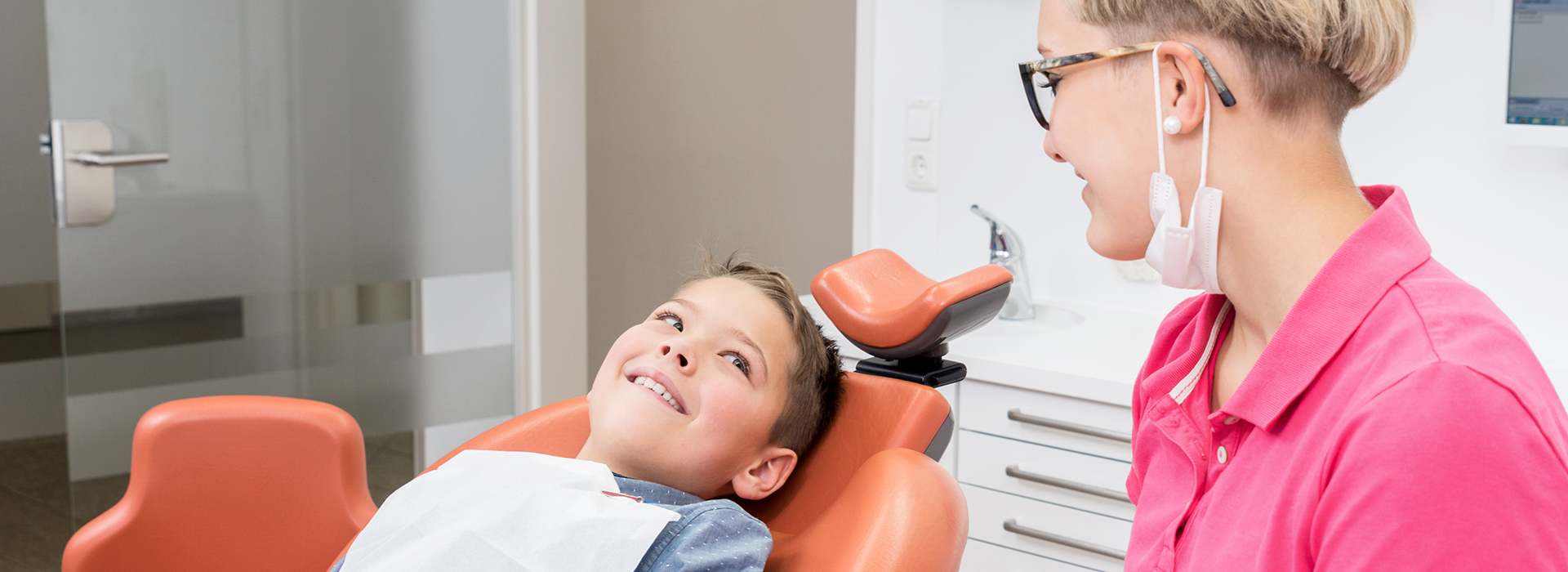 A dental patient smiling at the camera, with a dentist and hygienist in the background, sitting in a dental chair.