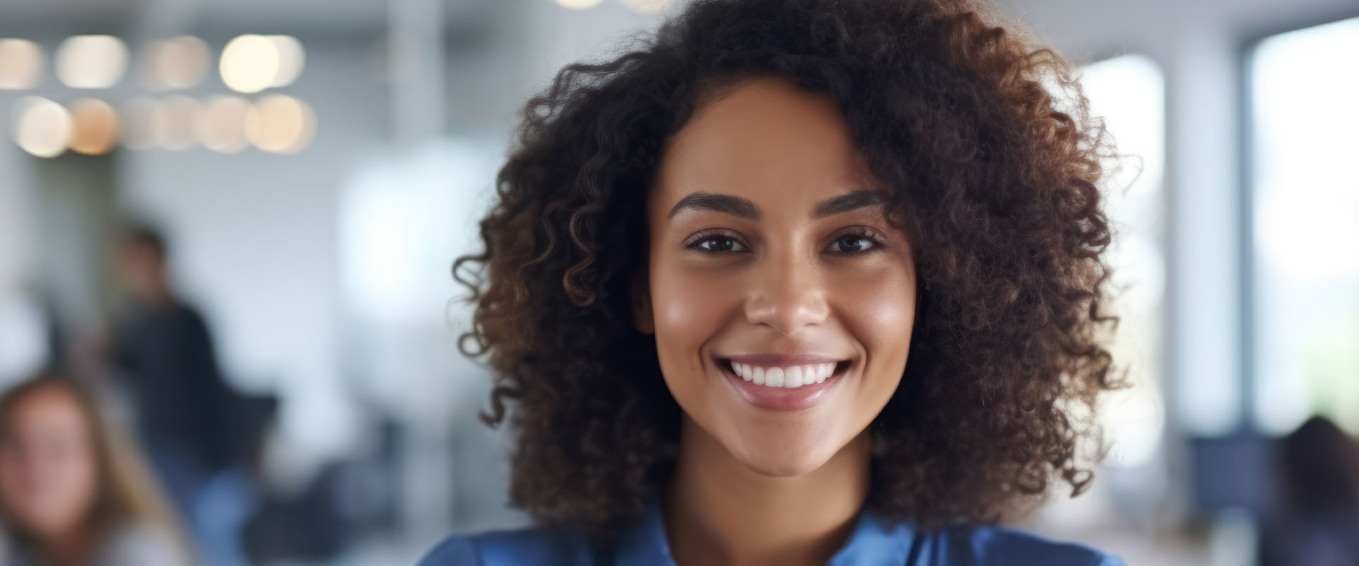 The image shows a person smiling at the camera, with a blurred background that suggests an indoor setting.