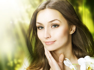 Woman with glowing skin, makeup, and a serene expression in front of a blurred background of greenery.