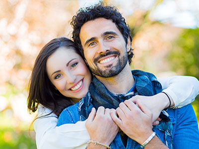 The image shows a man and a woman embracing each other, both smiling, with the man wearing a scarf around his neck. They are outdoors during daylight, and the setting appears to be a park or garden area.