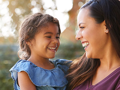 The image is a photograph of a woman and a young child, both smiling, with the woman holding the child. They appear to be outdoors during daylight.