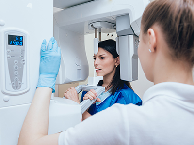 The image shows a woman in a lab coat standing next to a large, advanced piece of medical equipment, possibly an MRI machine, with another person who appears to be assisting or observing the equipment.