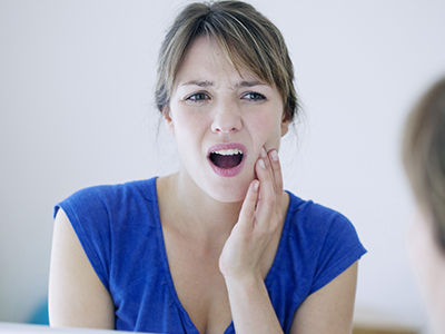 The image shows a woman with her mouth open, seemingly in pain or discomfort, holding her face and looking at her reflection in the mirror.