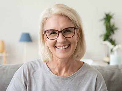 The image features a woman sitting on a couch, smiling at the camera. She has short blonde hair, is wearing glasses, and is dressed in a casual top with a light-colored background.