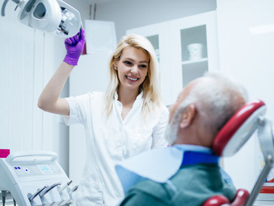 A dental hygienist assisting an elderly man in a dental chair, with the hygienist holding a mirror and smiling.