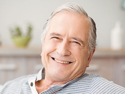 The image shows a smiling older man with grey hair, wearing glasses and a blue shirt, sitting in a relaxed posture with his hand on his chin.